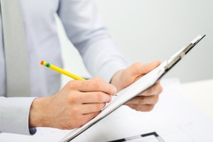 business, people and paperwork concept - close up of businessman hands writing to clipboard at office